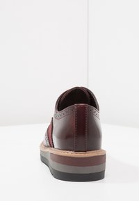 Chaussure en cuir bordeaux avec des détails de brogue, un talon bas et une semelle superposée en marron et gris. Design épuré avec un dos arrondi.