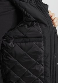 Sort polstret jakke med en quilted inderside, der har en sidelomme og en lynlås foran. Blød tekstur.