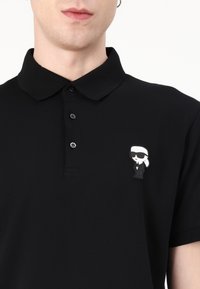 Fekete pamut póló klasszikus gallérral, három gombbal ellátva, bal mellen egy rajzfilmszerű kitűző részlettel.