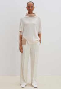 Strickpullover in sanftem Creme mit einem beigen Saum am unteren Ende, ausgestattet mit einem Cowl-Neck und kurzen Ärmeln, kombiniert mit weißen weiten Hosen.