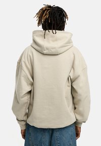 Beige Kapuzenpullover mit einer großen, verstellbaren Kordelzugkapuze, lässigen Schultern und gerippten Bündchen, von hinten vor einem schlichten Hintergrund dargestellt.