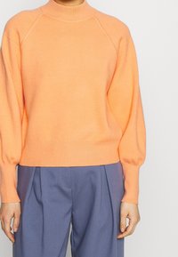 Suéter de punto naranja con cuello alto, corte relajado y puños acanalados. Presenta una textura suave y mangas raglán. Combinado con pantalones azul claro.