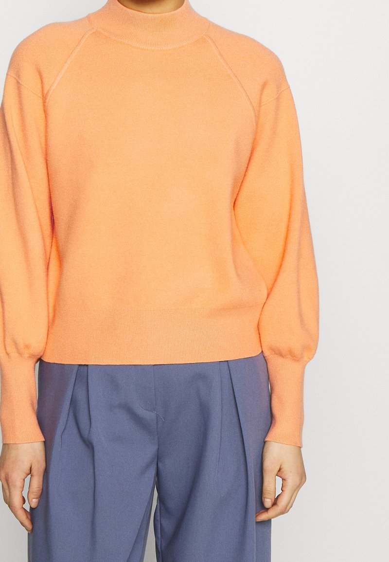 Suéter de punto naranja con cuello alto, corte relajado y puños acanalados. Presenta una textura suave y mangas raglán. Combinado con pantalones azul claro.