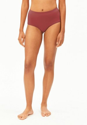 Femme debout pieds nus portant un sous-vêtement haut taille uni de couleur rouge foncé, les mains détendues le long du corps, sur fond blanc.