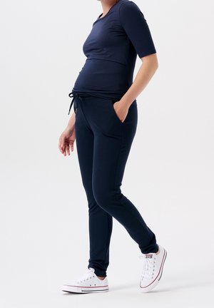 Femme enceinte portant un pantalon et un haut de maternité bleu marine, des baskets blanches, debout avec une main dans la poche sur un fond blanc.