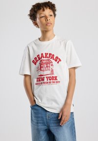White cotton t-shirt featuring a red graphic design of a bagel shop with text reading "Breakfast New York. Bagels as fresh as the city."