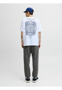 Hvid t-shirt med et blåt grafisk design på ryggen, grå bukser og navy kasket. Model iført sorte sko med hvide soles.