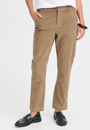 Personne portant un pantalon en velours marron clair, des mocassins noirs, un haut blanc et un bracelet noir, debout avec une main dans la poche.
