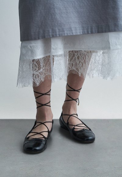 Ballerines en cuir noir avec des liens croisés autour de la cheville, ornées de détails perforés et d'un bout arrondi, assorties à une jupe blanche ornée de dentelle.