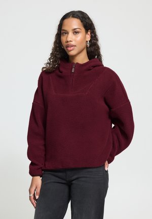 Femme aux cheveux bouclés portant des boucles d'oreilles créoles et un pull polaire bordeaux avec une fermeture éclair quart et un pantalon noir, debout devant un fond uni.
