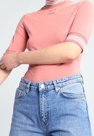 Osoba nosząca różową koszulkę Adidas z krótkim rękawem z białymi paskami na kołnierzu i rękawach oraz wysokim stanem niebieskie dżinsy.