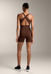Brun aktivkläder bodysuit med korsade axelremmar och öppen rygg, tillsammans med matchande shorts. Tyget är glatt med en figurnära silhuett.