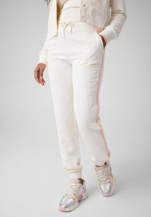Femme portant un pantalon de jogging blanc texturé avec des bandes latérales roses, des baskets crème avec des détails roses et beiges, et une veste assortie.