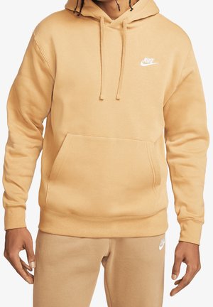 Person iført beige Nike-hættetrøje og matchende beige bukser med hvide Nike-logoer, stående foran en ensfarvet hvid baggrund.