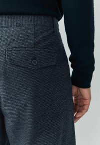 Zbliżenie na osobę w ciemnoszarych w kratę spodniach z zapinaną z tyłu kieszenią i czarnym długim rękawem, z ręką swobodnie opuszczoną wzdłuż ciała.