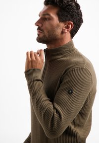 Geribbelde olijfgroene trui met een hoge kraag, voorzien van een rits en een hexagonaal logo patch op de linkermouw.