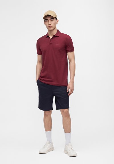 Homme portant une casquette beige, un polo bordeaux, un short marine, des chaussettes blanches et des baskets blanches, debout les mains dans les poches.