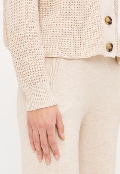 Beige knit cardigan with a textured pattern, featuring large brown buttons. Soft, matching beige pants with side pockets.
