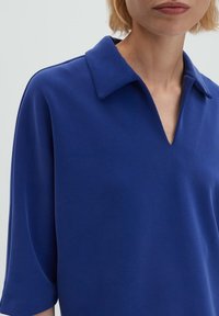 Femme portant une chemise bleue à large col en V, cadrée des épaules au milieu du torse, sur un fond clair uni.