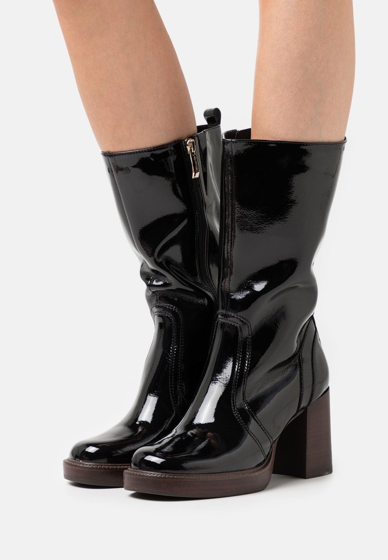 Bottes noires brillantes mi-mollet avec fermetures éclair latérales et talons carrés portées sur jambes nues sur un fond clair.