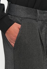 Grijze, gestructureerde broek met een glanzende tailleband. Zichtbare hand rustend in de zijzak, die de stofdetails en het contrast met de mouw van het shirt toont.