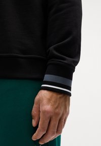 Felpa nera con polsini a coste caratterizzata da strisce grigie, nere e bianche; texture morbida; mano appoggiata su un tessuto verde sottostante.