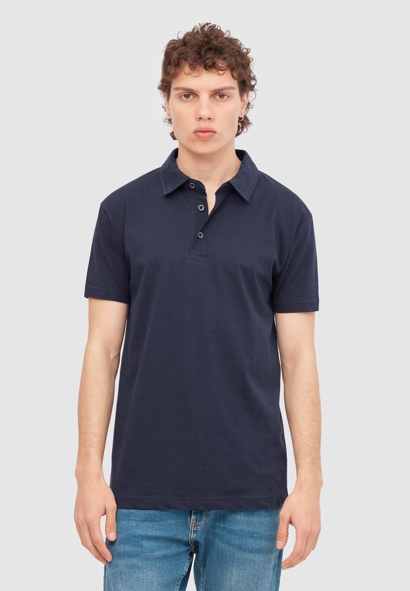 Camisa polo de color azul marino en algodón con cuello, mangas cortas, tapeta de tres botones y dobladillo recto, combinada con jeans de color azul claro.