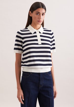 Kobieta o długich ciemnych włosach, ubrana w granatowo-białą koszulkę polo w poziome paski oraz ciemnoniebieskie spodnie z wysokim stanem, stojąca na jednolitym tle.