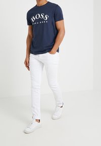 Tummansininen puuvillapaita, jossa on "BOSS"-logo, yhdistettynä valkoisiin slim-fit-farkkuihin ja valkoisiin lenkkareihin, esittelee rennoa ja modernia asua.