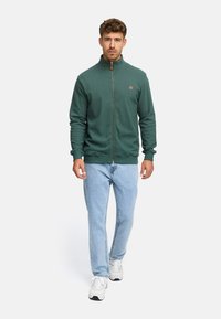 Mand der går fremad iført en grøn jakke med lynlås, lyseblå jeans og hvide sneakers på en lysegrå baggrund.
