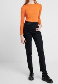 Person, die ein oranges Langarmshirt, hochgeschnittene schwarze Jeans und schwarze Schnürstiefel trägt, steht vor einem schlichten Hintergrund.