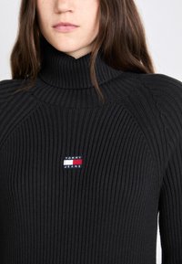Svart ribbad polo med turtleneck i stickad tyg. Har en liten "TOMMY JEANS" logotypapplikation i röd, vit och blå på bröstet.