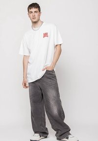 Young man wearing a loose white T-shirt with red text, baggy gray jeans, white sneakers, standing with one hand in pocket against a plain background.