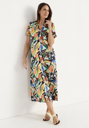 Kvinde i farverig midi-kjole med tropisk print og hvide sandaler, stående afslappet mod en hvid væg.