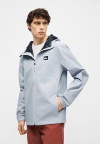 Νεαρός άνδρας που φοράει ανοιχτό γκρι αδιάβροχο μπουφάν με κουκούλα και λογότυπο, στέκεται με το ένα χέρ ι στην τσέπη και κοιτάζει προς το πλάι.