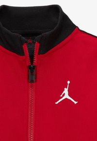 Punainen vetoketjullinen takki mustalla kauluksella, jossa on valkoinen Jumpman-logo edessä ja teksturoitu kangas. Vetoketju viimeistelee siistin ilmeen.