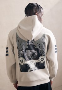 Beige hoodie med et stort grafisk print på ryggen. Viser en karakter med stråler bag sig. Tre sorte striber på hver ærme. Blød tekstur.