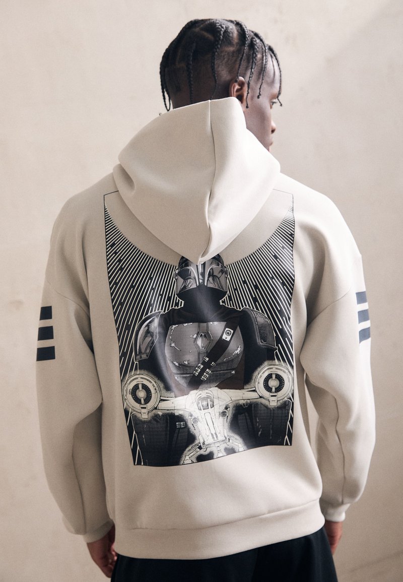 Beige hoodie med et stort grafisk print på ryggen. Viser en karakter med stråler bag sig. Tre sorte striber på hver ærme. Blød tekstur.