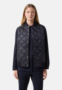 Marineblau gesteppte Weste mit Druckknöpfen und elastischen Akzenten am Saum. Über einem passenden marineblauen Oberteil getragen, das einen runden Ausschnitt hat.