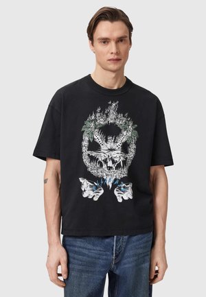 Junger Mann mit schwarzem T-Shirt, auf dem ein Grafikdesign mit einem brennenden Totenkopf, einem Marihuanablatt und blauen Flammen über offenen Händen abgebildet ist.