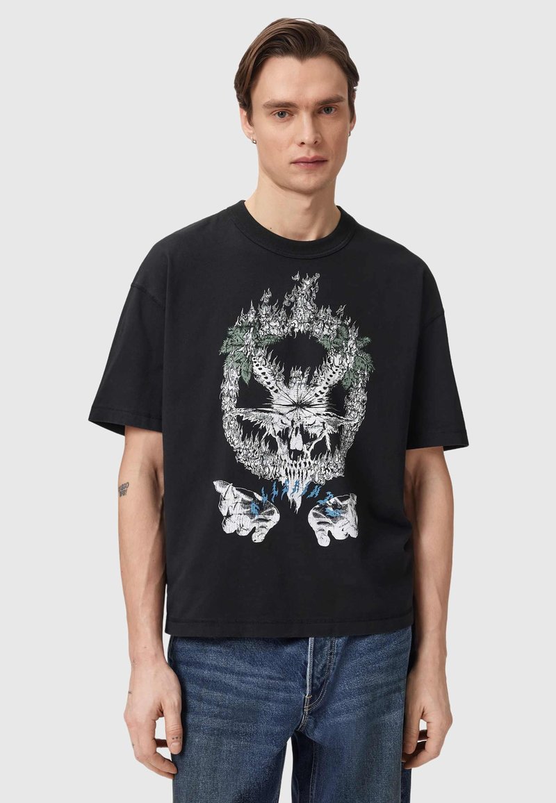 Νέος άνδρας που φοράει μαύρο μπλουζάκι με γραφικό από φλεγόμενο κρανίο, φύλλο μαριχουάνας και μπλε φλόγες πάνω από ανοικτά χέρια.