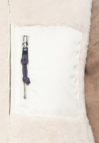 Detailný záber na vrecko bielej fleecovej bundy s vertikálnym strieborným zipsom a modro-červeným ťaháčikom na zips.