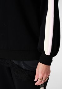 Zwarte sweater met een gestructureerde stof, met roze en witte strepen langs de mouwen. Voorzien van een kanten rand aan de onderkant.