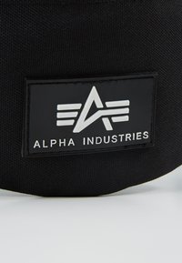 Svart stofflapp med teksten "ALPHA INDUSTRIES" og logo; har hevet design med vinklede former og horisontale detaljer i hvitt.