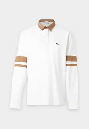 Ung mand iført en hvid langærmet skjorte med brune striber og krave, navy bukser og hvide sneakers, stående med hænderne i lommerne.