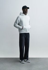 Grauer Kapuzenpullover mit Kängurutasche, kombiniert mit dunklen Denim-Hosen und schwarz-weißen Turnschuhen. Das Modell trägt eine schwarze Mütze.