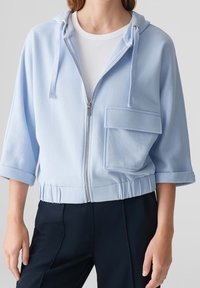 Osoba nosící světle modrou mikinu na zip s velkou přední kapsou a vyhrnutými tříčtvrtečními rukávy, v kombinaci s tmavými kalhotami.