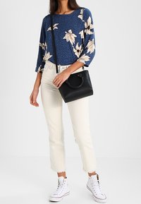 Sac à main noir avec une anse ronde, porté avec un haut bleu marine à motif floral et un jean blanc raccourci, assorti à des baskets blanches.