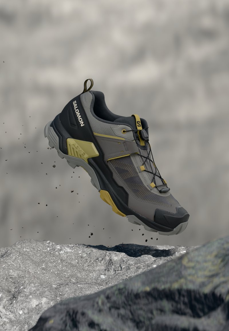 Scarpa da trail Salomon grigia e nera con accenti gialli sospesa sopra un terreno roccioso, mostrando la suola robusta e il sistema di allacciatura rapida.