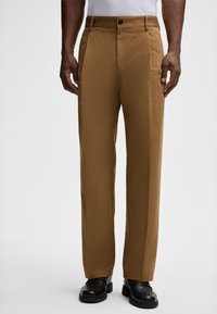 Pantaloni di cotone marroni con un design a gamba dritta, dotati di un unico bottone di chiusura e tasche laterali, indossati con scarpe nere.